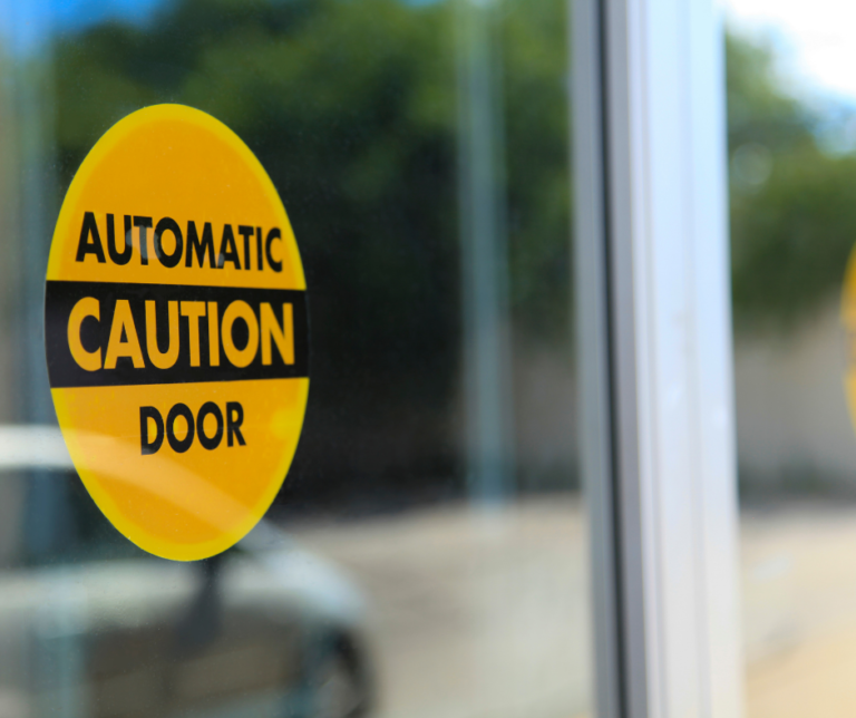 An image of a glass automatic door, with a yellow warning sticker that says “Caution Automatic Door”